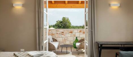 A bedroom with a bed, a small table, and a view of an outdoor balcony with a table and chairs.