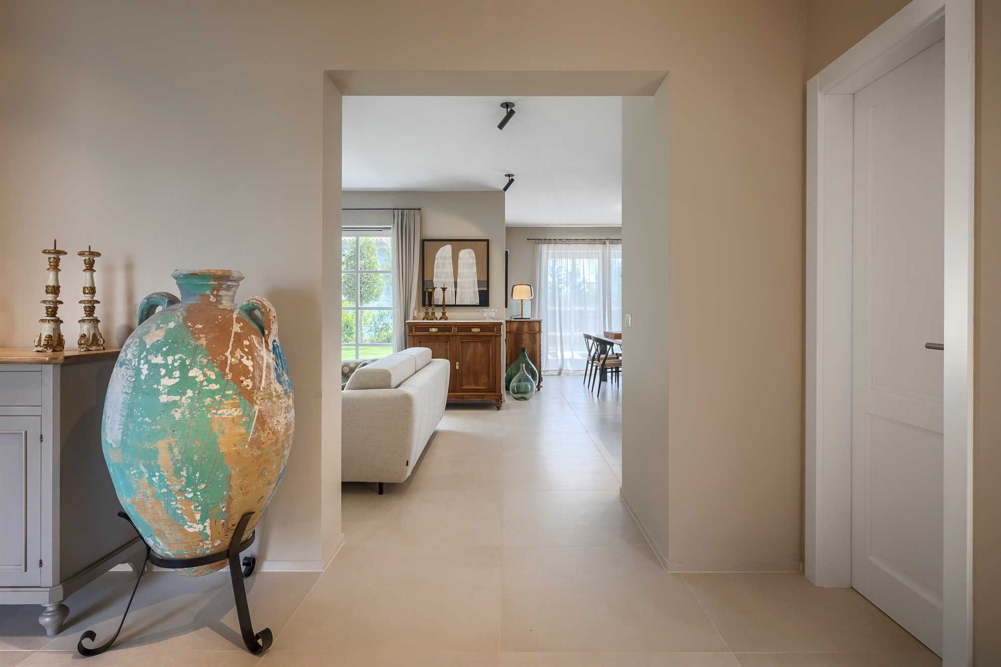 A hallway leading to a living room with a sofa, a dining table, and a lamp. There is a large decorative vase on wheels in the foreground.