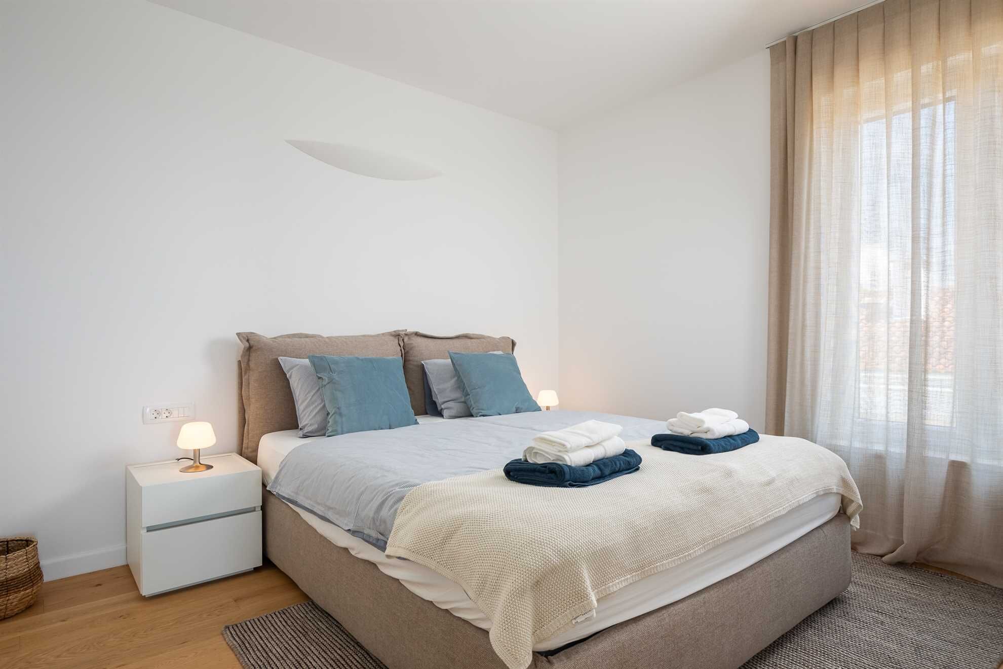 Ein ordentlich eingerichtetes Schlafzimmer mit einem Bett, Nachttisch, Lampe und einem Fenster mit Vorhängen.