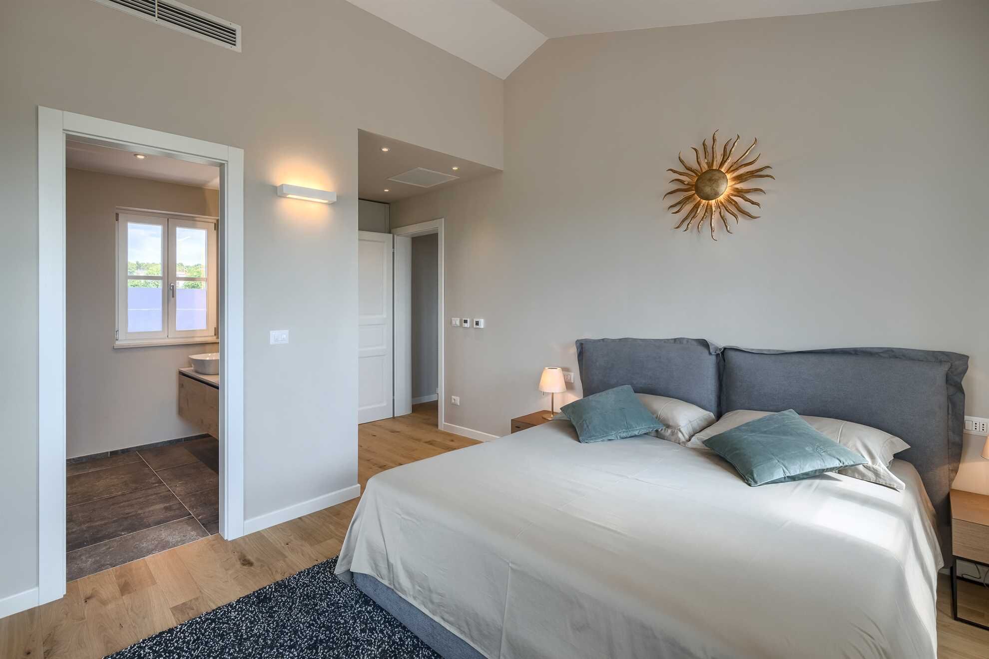 A bedroom with a bed, bedside tables, a nightstand, a wall-mounted light fixture, and a door leading to another room.