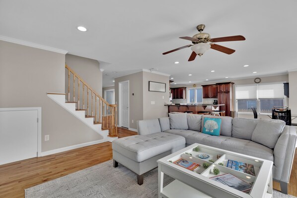 A modern living room with a grey sectional sofa, a white coffee table with magazines, a wooden staircase, and a ceiling fan.