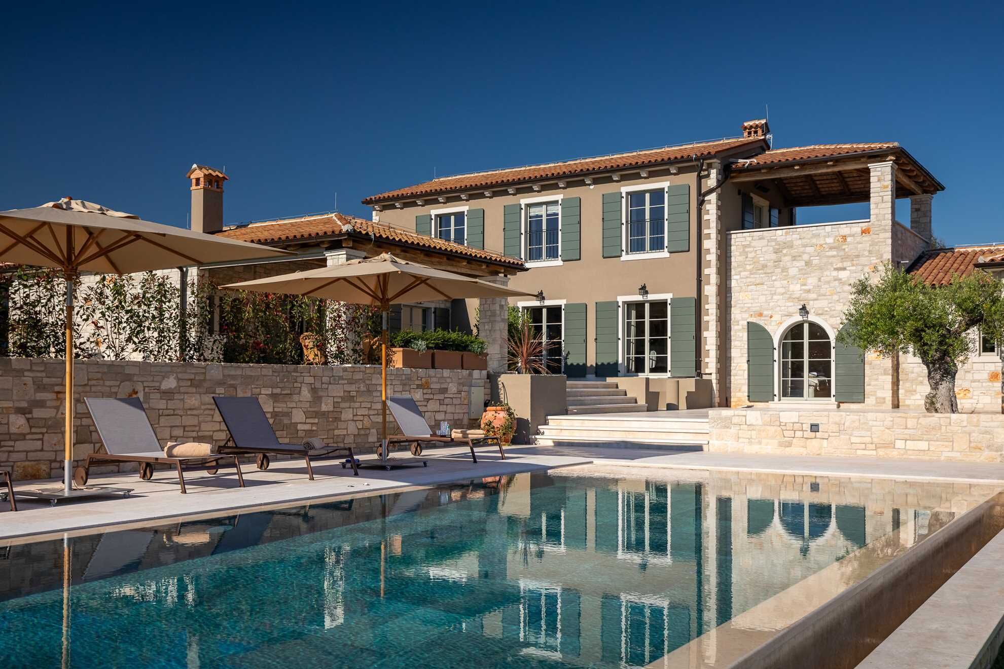 A pool area with lounge chairs and umbrellas, a stone wall, and a building with multiple windows and a tiled roof.