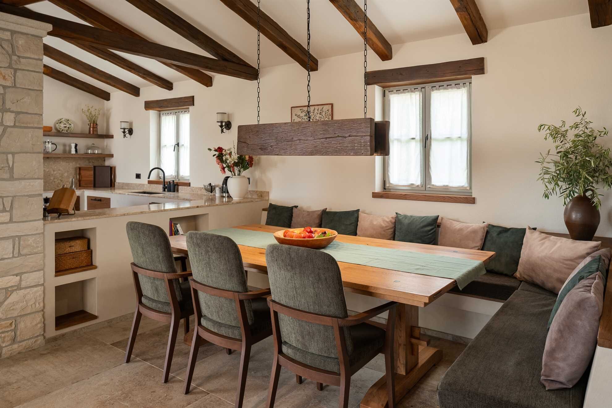 A cozy kitchen with a stone wall, wooden beams, a hanging light fixture, and a dining area with a wooden table and chairs.