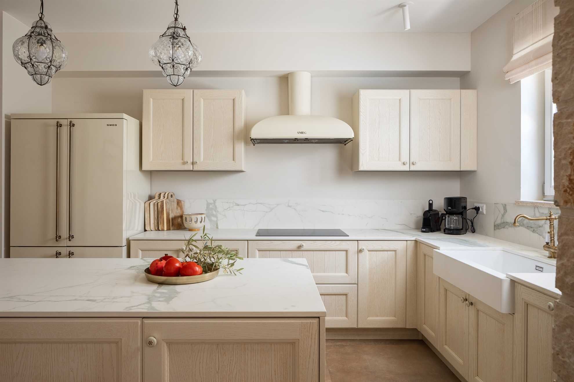 A kitchen with light wooden cabinets, a marble countertop, a central island with a bowl of tomatoes, a coffee maker, and a stove.