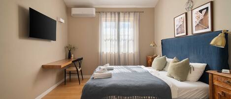 A bedroom with a bed, bedside tables, a desk, a chair, a TV mounted on the wall, and a window with curtains.