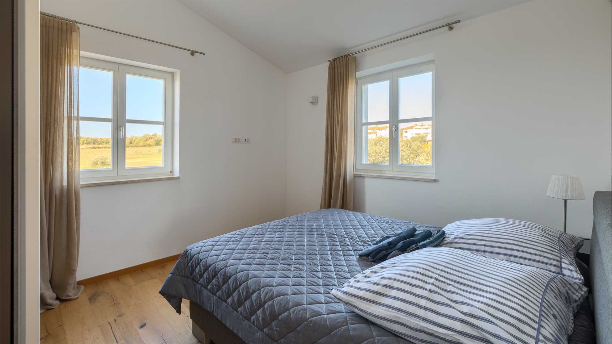 A bedroom with two windows, a bed with a quilted cover, a bedside lamp, and a view of a field.