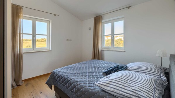 Un dormitorio con dos ventanas, una cama con cubrecama acolchado, una lámpara de noche y vistas a un campo.