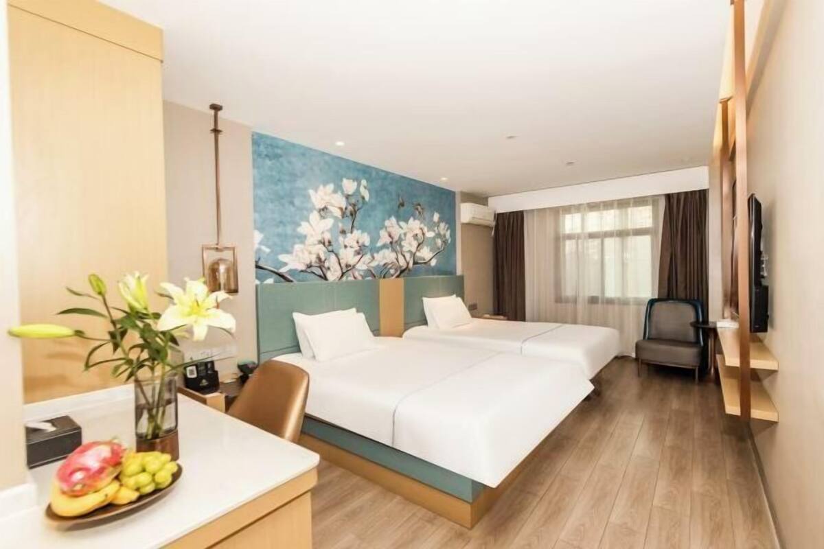 A hotel room with two beds, a desk with a vase of flowers, a painting on the wall, and a television.