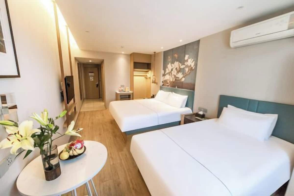 A hotel room with two beds, a small table with a vase of flowers, a wall-mounted air conditioner, and a painting.
