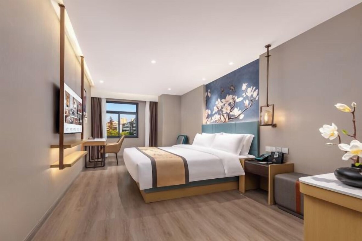 A hotel room with a bed, bedside table, desk, chair, and a painting on the wall.