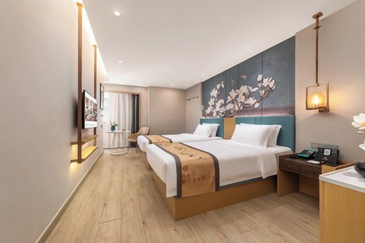 A hotel room with two beds, a desk, a chair, a painting on the wall, and a nightstand with a lamp.