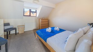 Een slaapkamer met een schuine kap, een bed met een blauwe deken, een dressoir, een stoel, een radiator en een raam met uitzicht op gebouwen.