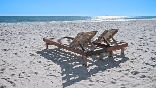 Une chaise longue en bois sur une plage de sable, parsemée d’empreintes, face à l’océan.