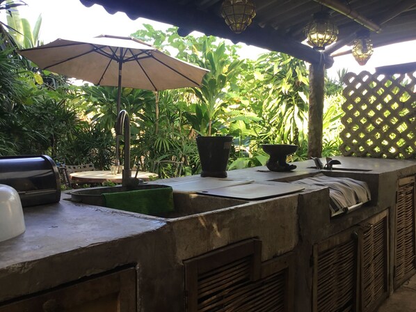 An outdoor bar area with a concrete countertop, a grill, a large umbrella, and lush greenery in the background.