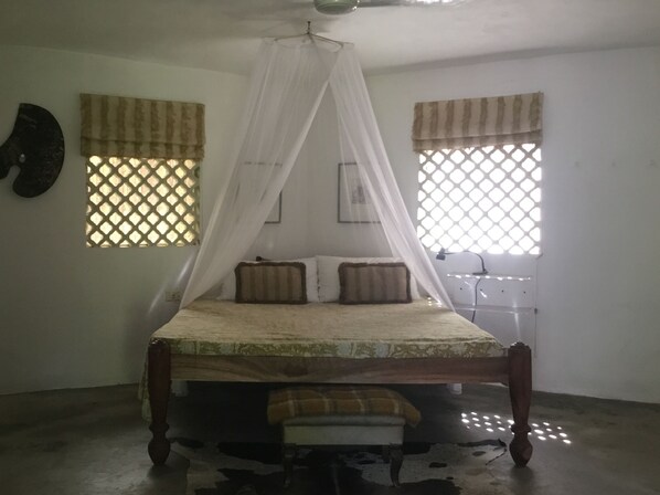 A bedroom with a canopy bed, two windows with patterned curtains, a bedside table, and a wall-mounted clock.