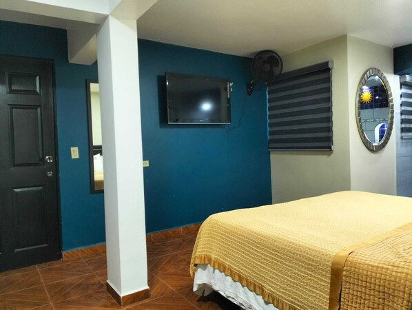 A bedroom with a bed, a television mounted on the wall, a window with blinds, and a mirror.