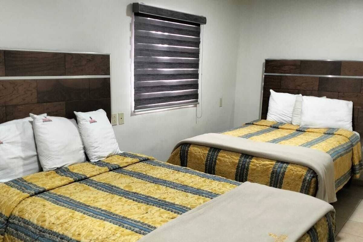 Ein Hotelzimmer mit zwei Betten, jeweils mit Kopfteil, einem Fenster mit Jalousien und einem Fliesenboden.