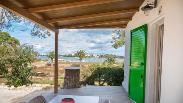 Un área de estar al aire libre techada, con una mesa y sillas, una puerta verde y vistas al mar y al paisaje.