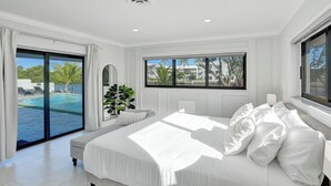 A bedroom with a large bed, a view of a swimming pool, and a window overlooking palm trees.