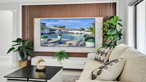 A modern living room with a large TV displaying a poolside scene, a white sofa, a coffee table, and decorative plants.