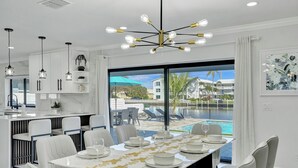A modern dining area with a large table set for six, white chairs, and a view of a swimming pool and buildings.