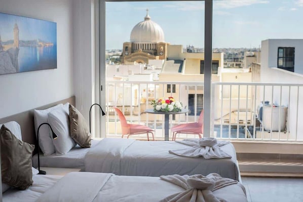 A hotel room with a bed, a small table with flowers, a balcony with chairs, and a painting on the wall.
