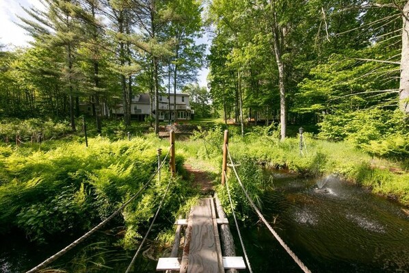 A wooden suspension bridge over a stream in a forested area with dense greenery and a house in the background.