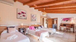 A hotel room with two beds, a desk, and a chair. There are paintings on the walls and a ceiling fan. - Villa es Rafal by Mallorca North Villas  (Santa Margalida)