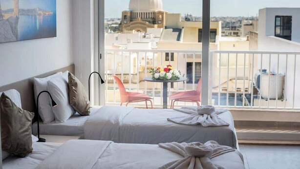 Quarto de hotel com cama, uma pequena mesa com flores, varanda com cadeiras e um quadro na parede.