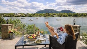 Une personne se détend sur une chaise près d’une table, avec vue sur un lac et des bateaux, entourée de verdure et avec une montagne en arrière-plan.
