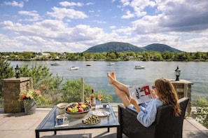 A person relaxing on a chair by a table with a view of a lake and boats, surrounded by greenery and a mountain in the background.