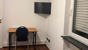 A small, compact room with a wooden desk, a chair, a bed, a mounted TV, and a window with blinds. - NR 6 ground floor 3 ZKB for families and companies (Fuldatal)