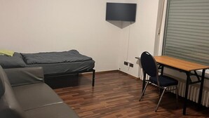 A compact room with a sofa, a small table with a chair, a flat-screen TV mounted on the wall, and a window with blinds. - NR 6 ground floor 3 ZKB for families and companies (Fuldatal)
