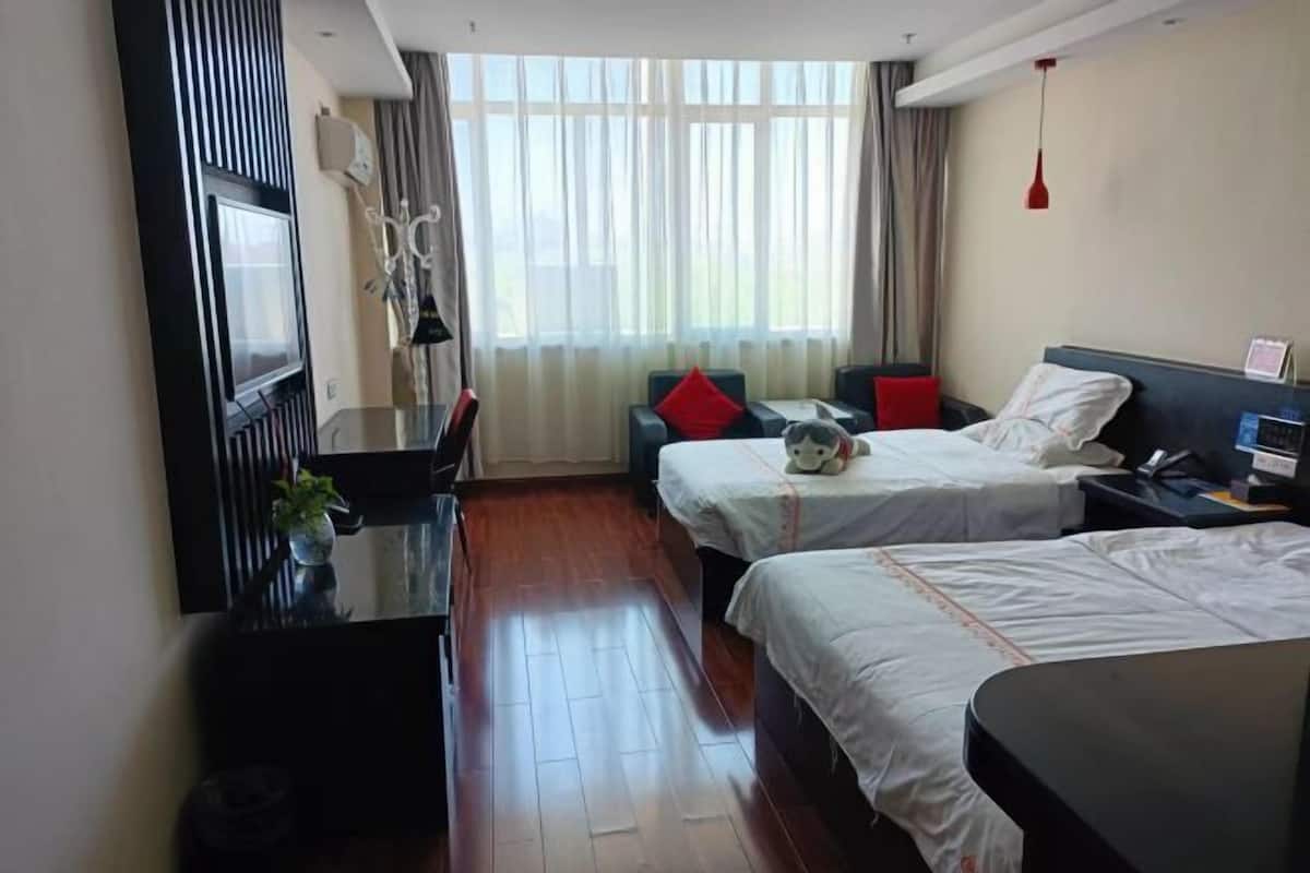 A hotel room with two beds, a desk, a TV, and a large window with curtains.