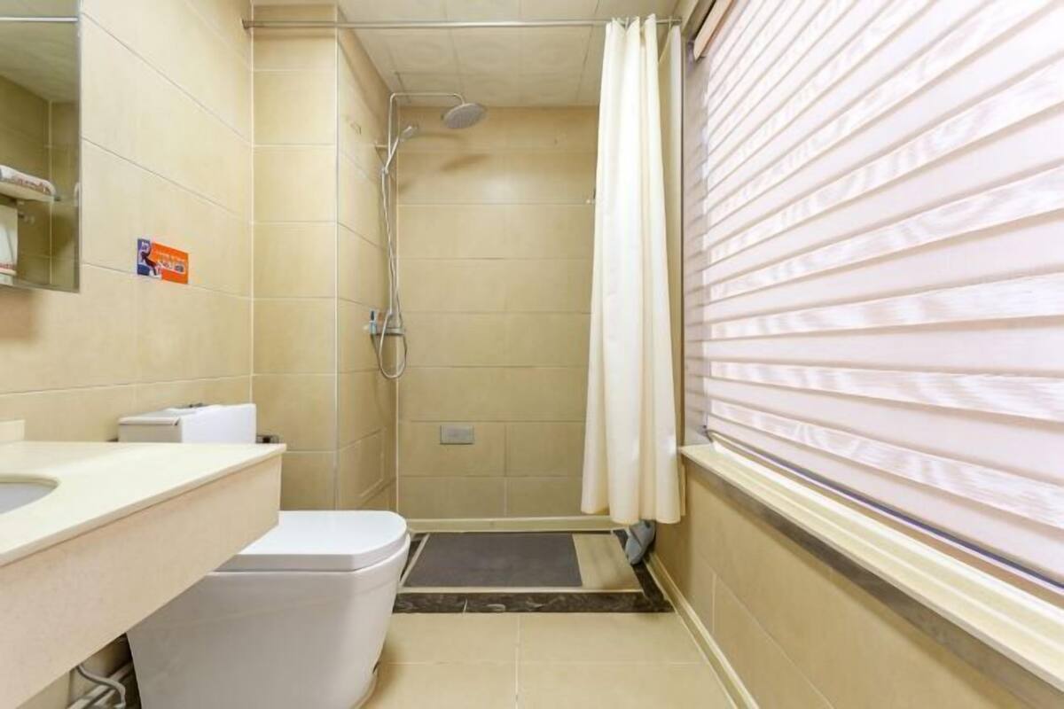A bathroom with a toilet, sink, mirror, shower, and window with blinds.