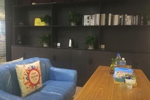 Una habitación con un sofá azul, una mesa de madera con una caja y una botella, y una estantería con libros y plantas.