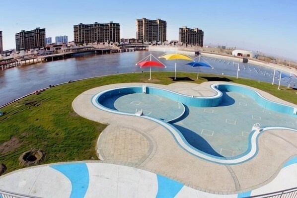A waterfront park with a winding pool, umbrellas, and a view of the city skyline.