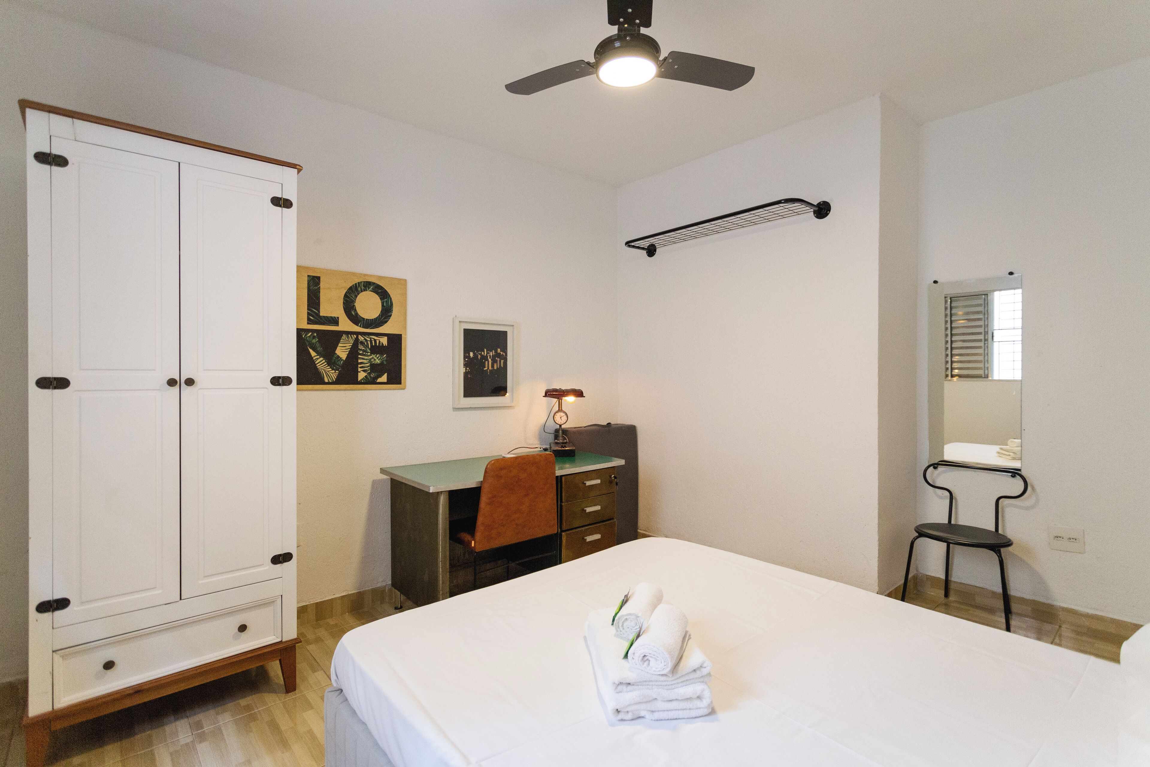 A hotel room with a bed, a desk, a wardrobe, a mirror, and a ceiling fan.