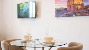 Een eetgedeelte met een ronde glazen tafel, twee stoelen en een aan de muur bevestigde televisie.