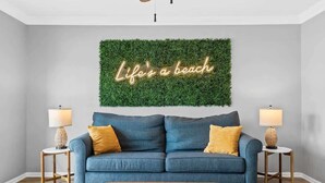 Una sala de estar con un sofá azul, dos mesitas de noche con lámparas, un reposapiés marrón y un letrero de neón en la pared.
