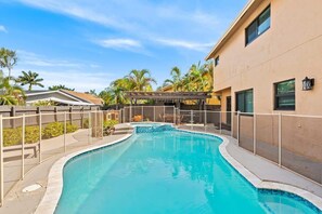 En swimmingpool med en buet kant, omgivet af et hegn, ved siden af en bygning med en pergola.