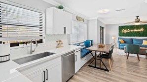 Una cocina moderna con lavavajillas incorporado, una zona de comedor con mesa y sillas de madera, y un espacio de sala con sofá azul y un letrero de neón.