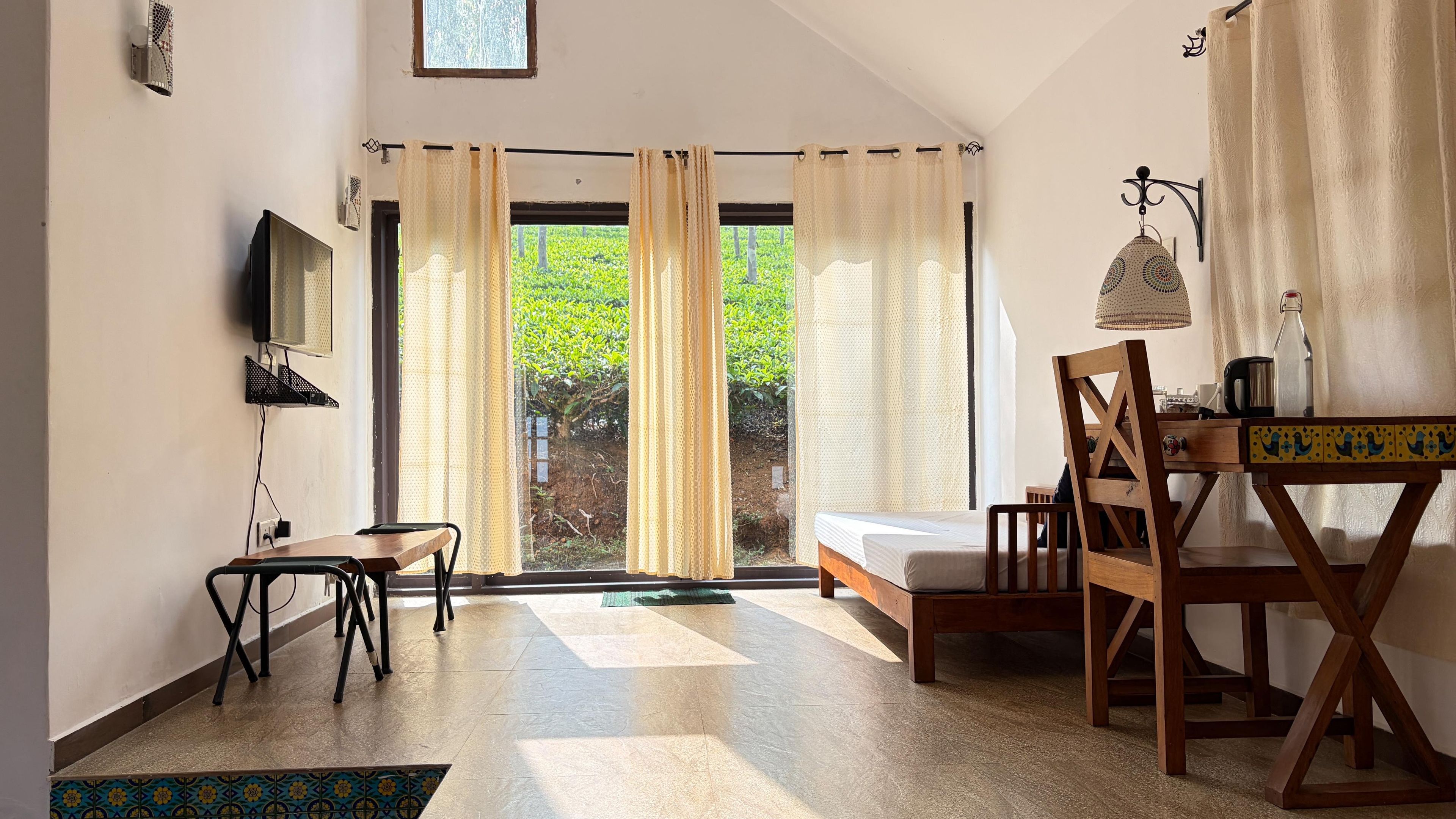 A room with a bed, a desk, a chair, a TV, and a view of greenery outside.