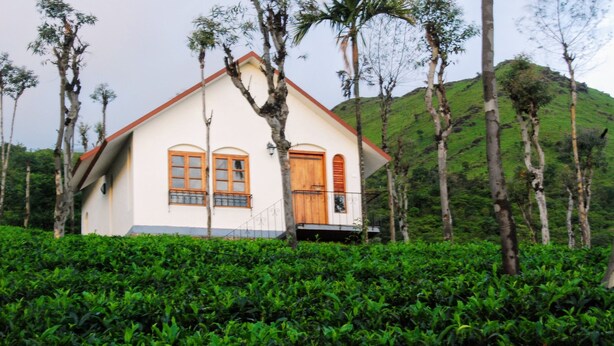 A white building with a red roof, surrounded by lush green tea bushes and tall trees. - Tea Terrace Vythiri Resort (Vythiri)