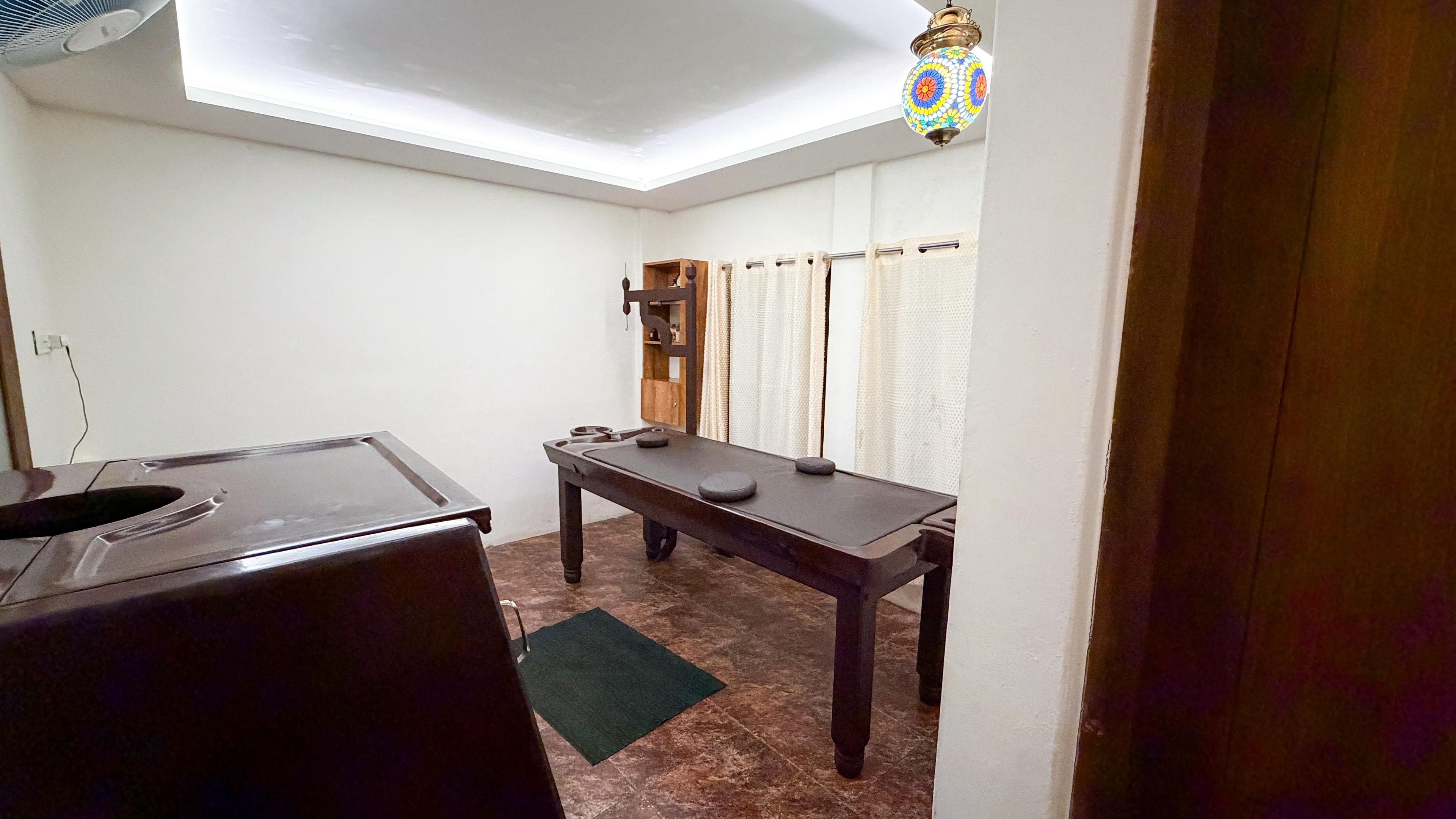 Ein Zimmer mit Massagetisch, einer kleinen Holzkabine, Deckenventilator und einer dekorativen Pendelleuchte.