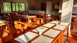 A dining area with wooden tables and chairs, large windows with a view of greenery, and a television on the wall. - Tea Terrace Vythiri Resort (Vythiri)