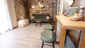 A cozy living room with a stone wall, a green sofa, two green stools, a wooden coffee table, and a kitchen area with a coffee machine and a basket of bread. - Lo Raconet dárnes, a rural apartment in the historical center of Arnes. (Arnes)