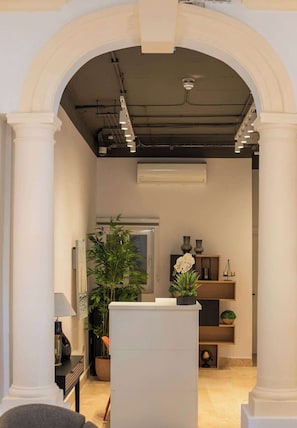 An interior space with a reception desk, a potted plant, a wall-mounted air conditioner, and a shelf with decorative items.