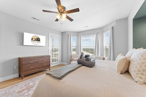 Une chambre Ă coucher comprenant un lit, un ventilateur de plafond, une tĂ©lĂ©vision, une commode et une fenĂȘtre avec des rideaux.