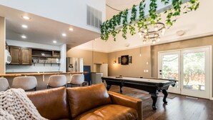 A living room with a brown leather sofa, a pool table, and a kitchen area with cabinets and a window. - Treetop Castle *Sleeps 16* (Boise)